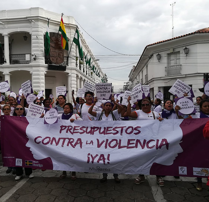 Marcha de mujeres frente a un edificio colonial blanco con la bandera boliviana. Sostienen una pancarta que dice «Presupuestos contra la violencia ¡YA!» y carteles con la consigna «Vivas nos queremos».