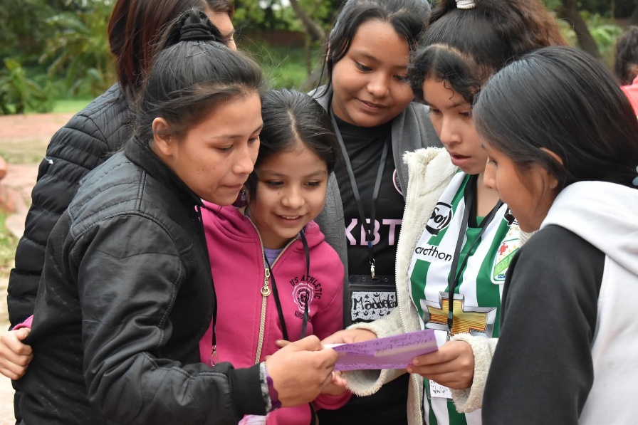 Una mujer, una adolescente y una niña leen juntas, junto a otras compañeras, una nota escrita a mano