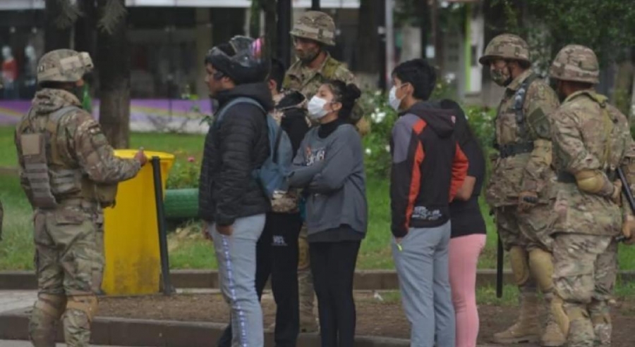 Militares con uniforme de camuflaje, casco y armas rodean a un grupo de jóvenes con barbijo en una plaza con césped y canteros.