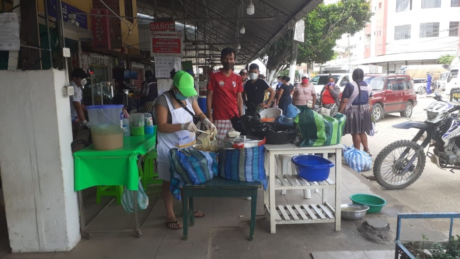 Puesto de comida callejero bajo un techo de chapa. Una mujer con delantal, gorra y barbijo sirve alimento en bolsas; alrededor hay personas con barbijo, bolsas de mercado y una motocicleta.