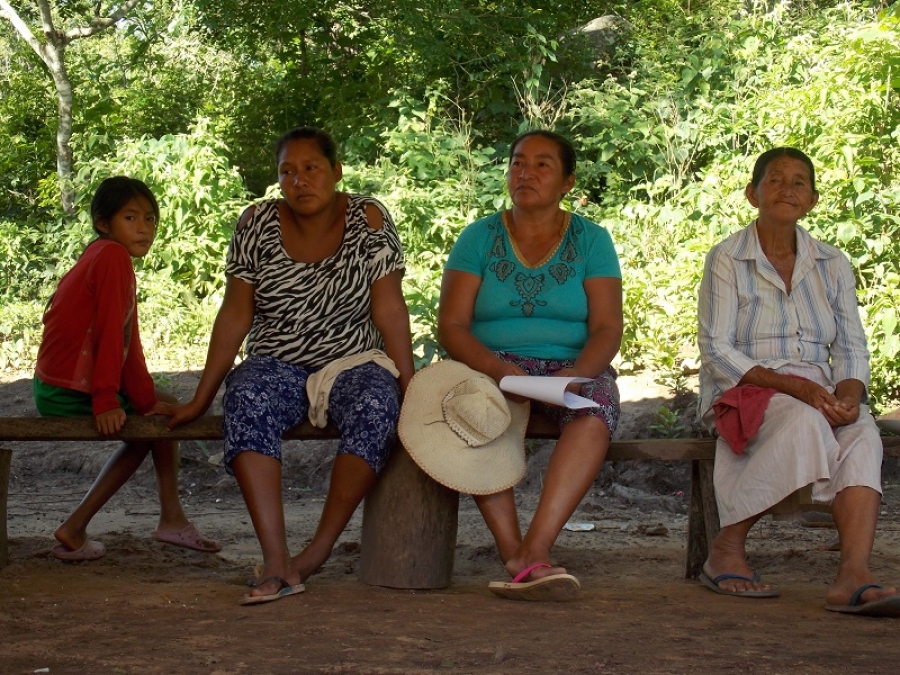 Tres mujeres y una niña sentadas en un banco de madera a la sombra de los árboles, en un entorno rural.