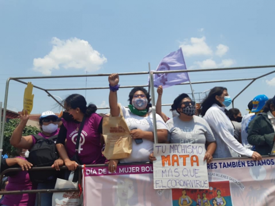 Movilización sobre una plataforma: mujeres con barbijo, una con el puño en alto y otra ondeando una bandera violeta. Una sostiene un cartel escrito a mano que dice «Tu machismo mata más que el coronavirus». Abajo cuelga una pancarta por el Día de la Mujer.