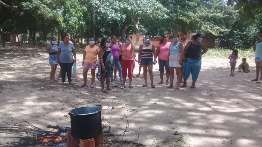 Un grupo de mujeres con barbijo, de pie en fila sobre un descampado de tierra entre árboles. En primer plano, una olla grande puesta sobre un fuego de leña.